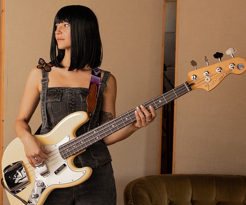 Woman holding a vintage white Fender Jazz Bass guitar, standing in a cozy indoor studio setting.