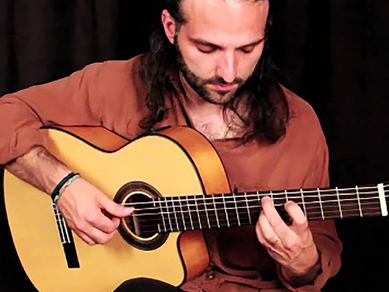 Man with long dark hair and beard playing a flamenco or classical guitar with a spruce top in a close-up performance shot