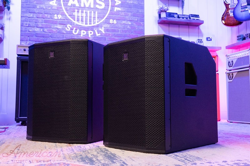 Alt tag: Wall-mounted white indoor speaker with a perforated grille, installed below a slatted wood ceiling.