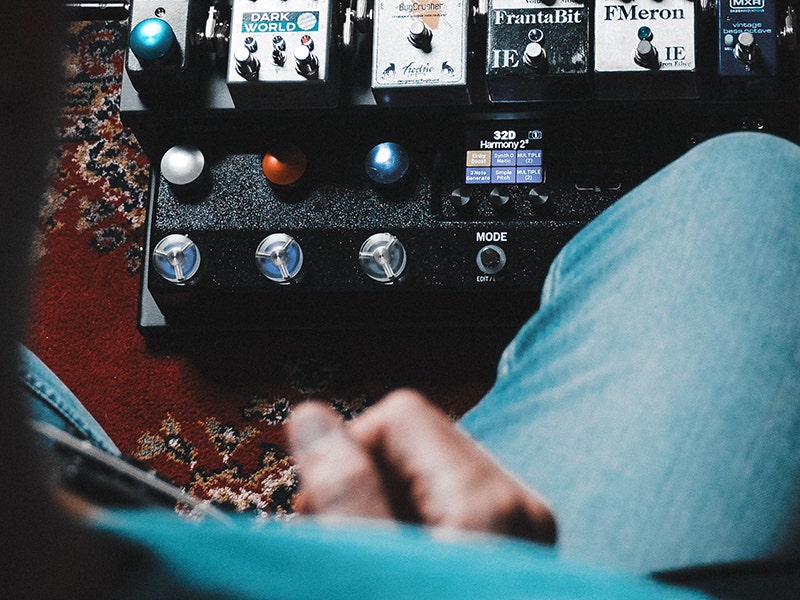Overhead view of a seated guitarist adjusting a pedalboard loaded with various guitar effects pedals.