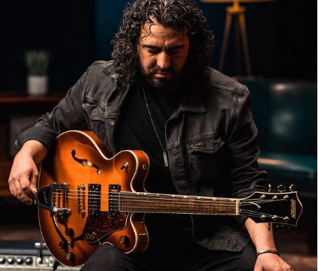A male musician sitting on a stool with a hollow-body Gretsch guitar.