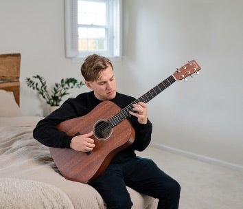 A man sitting on a bed plays a Martin X Series acoustic guitar.