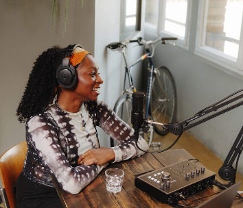 A smiling woman wearing Audio-Technica headphones sits at a wooden table with a microphone and audio mixer.